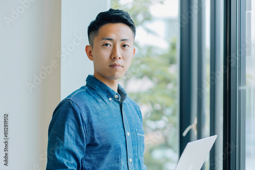 Young Asian man holding a laptop in his hand, standing at a window and looking to the camera with a confident expression while working on a digital marketing project or writin photo