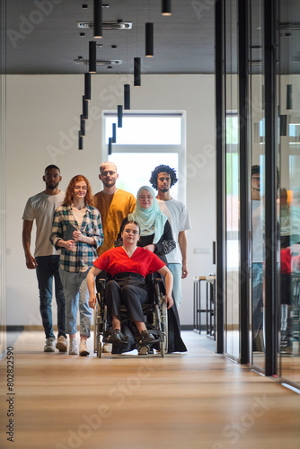 A diverse group of young business people walking a corridor in the glass-enclosed office of a modern startup, including a person in a wheelchair and a woman wearing a hijab