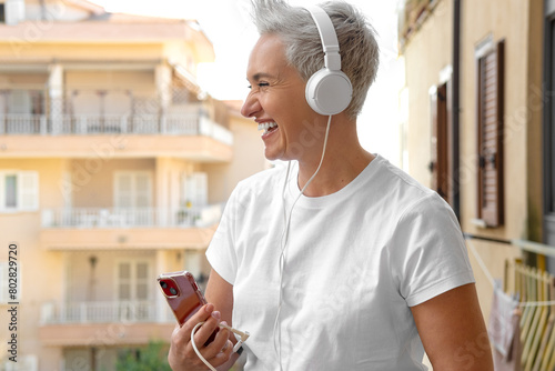 Middle Aged Woman With Short Haircut Listening To Music With Her Smartphone. Audiobook or Podcast Concept. Active Life Position. Older Adults and Technology Use. Laughing