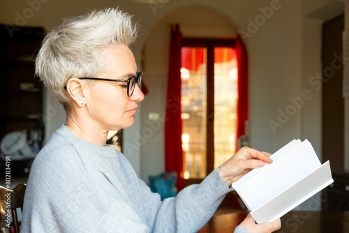 Middle-Aged Mature Businesswoman Holding And Looking At An Empty White Paper Book In The Home Background. Book Templates And Mockup. Personal Development After 40. Personal Growth Or Change After 45.