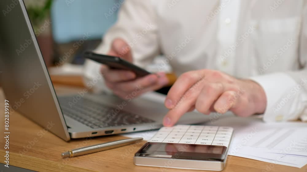 Man Working in Office, Using Laptop Computer and Calculator Placed on Desk, Holding Mobile Cell Phone close up. Male professional using computer modern notebook sitting at home office desk Man Working in Office, Using Laptop Computer and Calculator Placed on Desk, Holding Mobile Cell Phone close up. Male professional using computer modern notebook sitting at home office desk