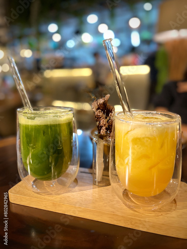 A glass of cold orange juice with ice and a glass of healthy green juice with glass stirs at a restaurant setting with blurry background