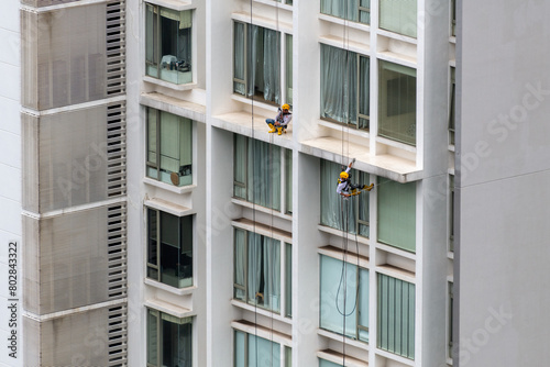 Two men at the exterior of a high-rise building. One working at height cleaning window and the other one sleeping on the job