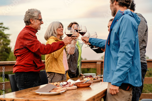 A group of mature friends - toasting with wine glasses at an outdoor table, surrounded by nature at sunset - joyful, festive gathering. © Lomb
