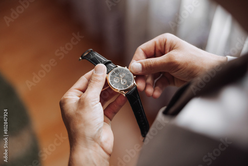 A man is holding a watch and looking at it. The watch is black and gold. The man is wearing a suit