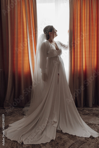 A woman in a wedding dress stands in front of a window