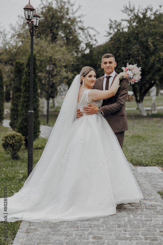 A bride and groom are posing for a picture in a park. The bride is wearing a white dress and the groom is wearing a suit. They are holding hands and the bride is holding a bouquet A bride and groom are posing for a picture in a park. The bride is wearing a white dress and the groom is wearing a suit. They are holding hands and the bride is holding a bouquet