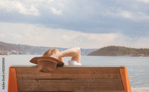 Young blond-haired woman with brown hat sits on her back on wooden bench realxing and watching sea bay summer time vintage style © Wioletta