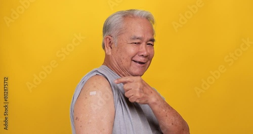 Asian old man showing adhesive plaster bandage on shoulder after vaccination. Isolated on yellow background in the studio. photo