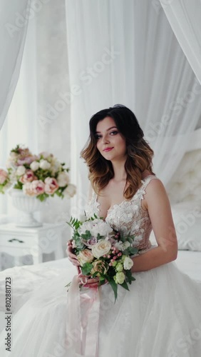 Beautiful young model in a white lace dress sits on a white bed. Gorgeous bride in wedding dress with flowers is sitting in the light bedroom. Vertical video