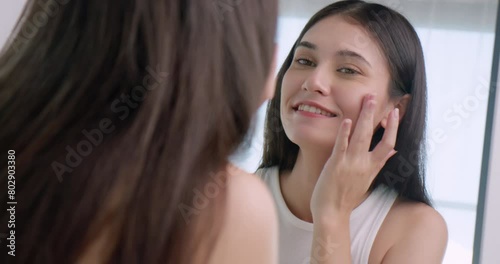 Beautiful young woman applies moisturizer to her face while standing in front of a mirror. photo