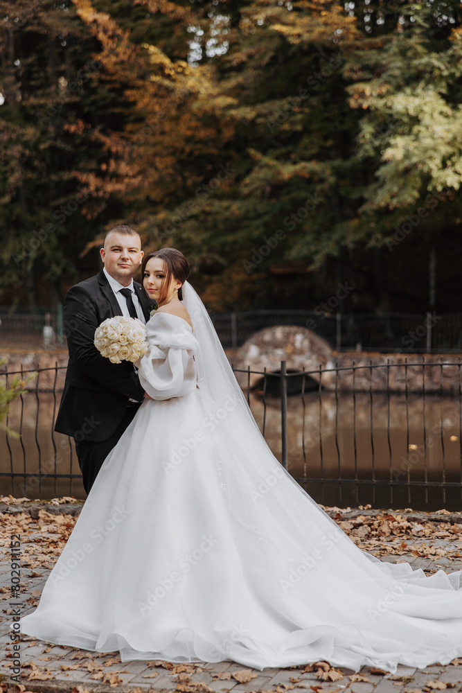 A bride and groom pose for a picture in front of a bridge. The bride is wearing a white dress and the groom is wearing a suit