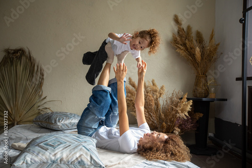 Smiling young Caucasian mom lying on comfortable bed have fun engaged in funny game activity with small daughter, overjoyed mother and little girl child playing together in cozy bedroom at home