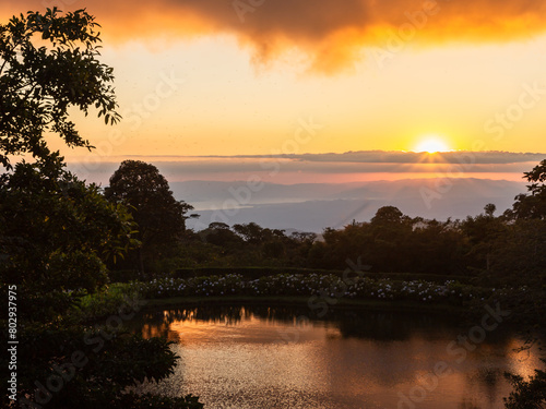 High angle view of small lake in hilly terrain with reflection of the setting sun and the Pacific Ocean in the background, Monteverde, Costa Rica