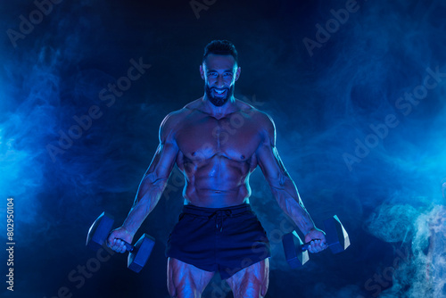 Sporty fit man athlete with dumbbells make fitness exercises on blue neon background. Download cover for music collection for fitness classes. Sports recreation. © Mike Orlov