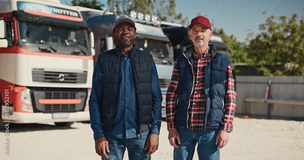 Obraz premium Two mixed-race coworkers standing before large trucks outside. Mature drivers in hats and jackets positively looking into camera. Getting ready to traveling. Working together in delivery company.