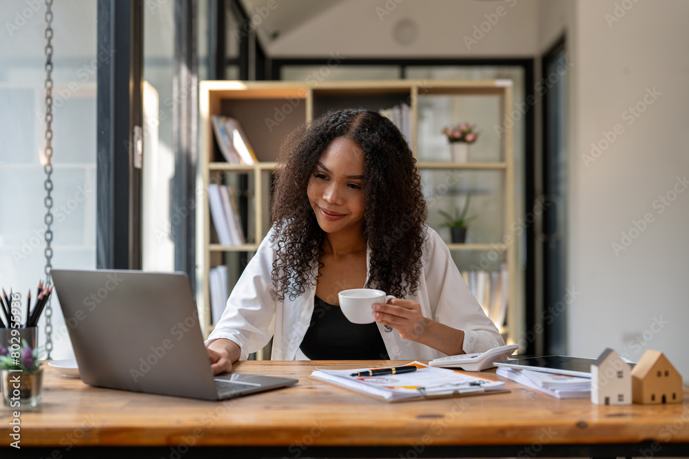 A woman is sitting at a desk with a laptop and a cup of coffee. She is smiling and she is enjoying her coffee A woman is sitting at a desk with a laptop and a cup of coffee. She is smiling and she is enjoying her coffee