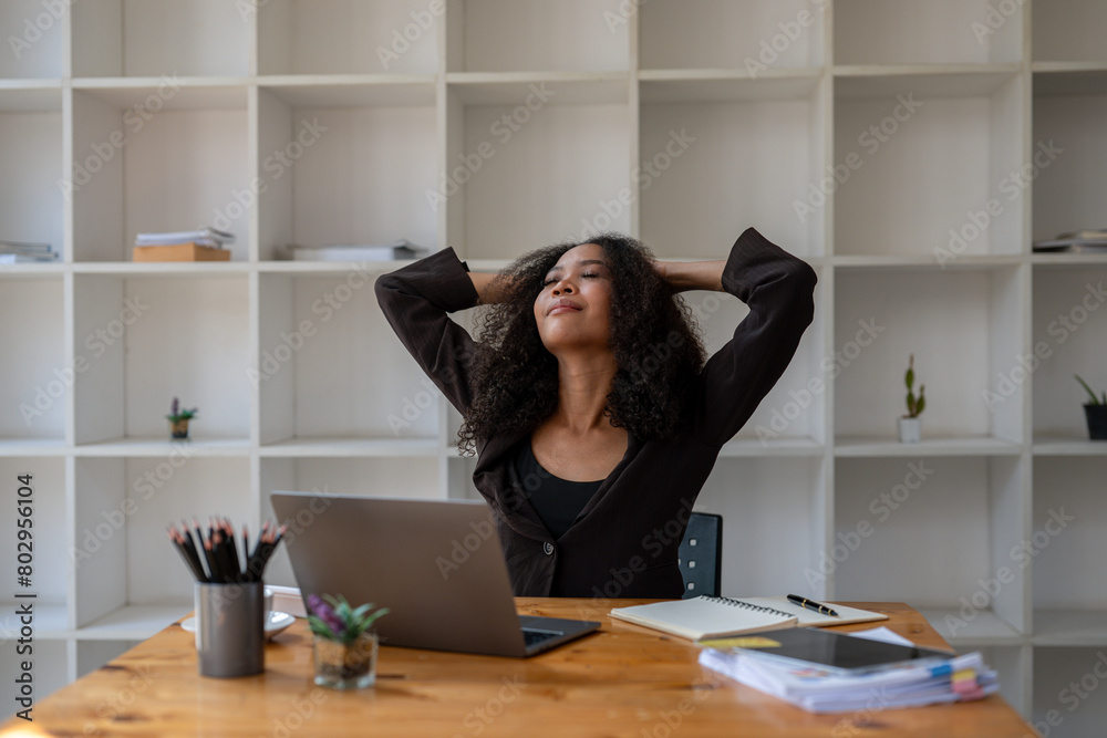 © Wasana - A woman is sitting at a desk with a laptop and a notebook. She is looking at the laptop and she is in a relaxed state © Wasana - A woman is sitting at a desk with a laptop and a notebook. She is looking at the laptop and she is in a relaxed state