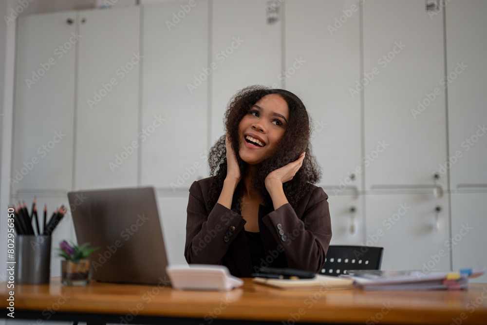 A woman with curly hair is sitting at a desk with a laptop and smiling. She is looking at the camera and she is happy A woman with curly hair is sitting at a desk with a laptop and smiling. She is looking at the camera and she is happy