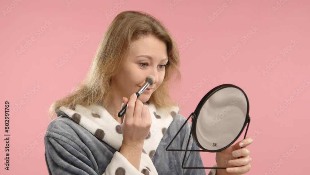 Captured against a soft pink background, a young woman applies makeup with precision, using a brush while gazing into a hand mirror, dressed in a polka dot robe. Captured against a soft pink background, a young woman applies makeup with precision, using a brush while gazing into a hand mirror, dressed in a polka dot robe.