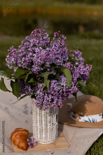 Bouquet of lilacs in a wicker vase, croissant and hat. Spring Picnic