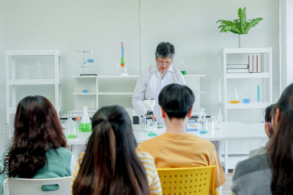 Students' learning in science. Asian educator in lab coat demonstrating microscope usage to attentive young students in science class with various lab glassware on white desks. Students' learning in science. Asian educator in lab coat demonstrating microscope usage to attentive young students in science class with various lab glassware on white desks.