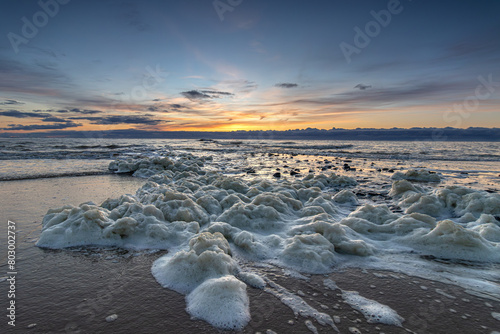 Sea foam washes up on the beach of Den Helder near a pier during a colorful sunset. Foam on seawater, formed by dead slime algae, washes up on the beach during a colorful sunset. photo