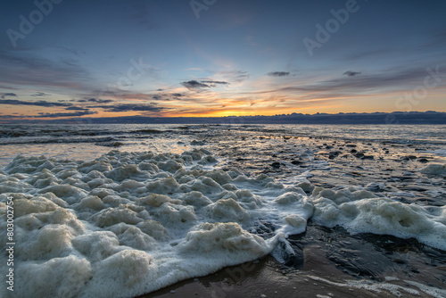 Sea foam washes up on the beach of Den Helder near a pier during a colorful sunset. Foam on seawater, formed by dead slime algae, washes up on the beach during a colorful sunset.  photo