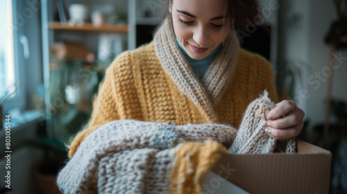 Close-up: The young woman owner entrepreneur delicately wraps a soft, pastel-colored sweater and carefully places it in a shipping box, her computer screen displaying incoming onli photo