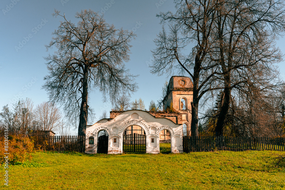The main entrance to the territory of the temple. An abandoned, crumbling church in a green meadow, behind a white gate.