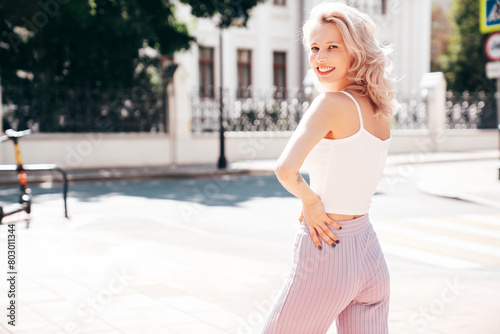 Young beautiful smiling blond hipster woman in trendy summer clothes. Carefree female posing in the street at sunny day. Positive model outdoors at sunset. Cheerful and happy