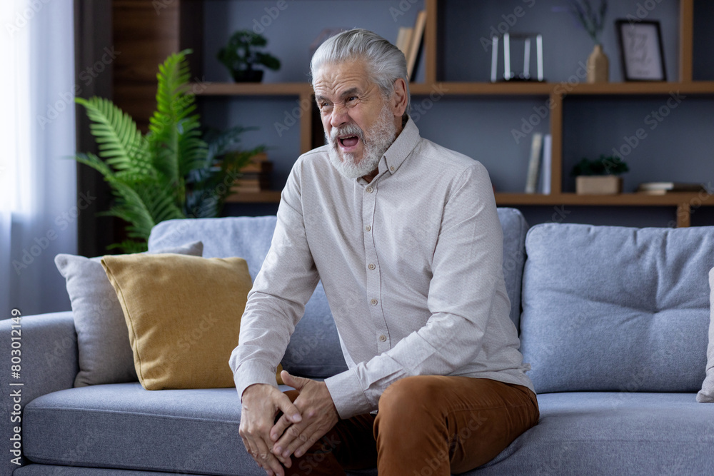 Grimacing from severe pain, an older gray-haired man sits on the sofa at home and holds his knee with his hand Grimacing from severe pain, an older gray-haired man sits on the sofa at home and holds his knee with his hand