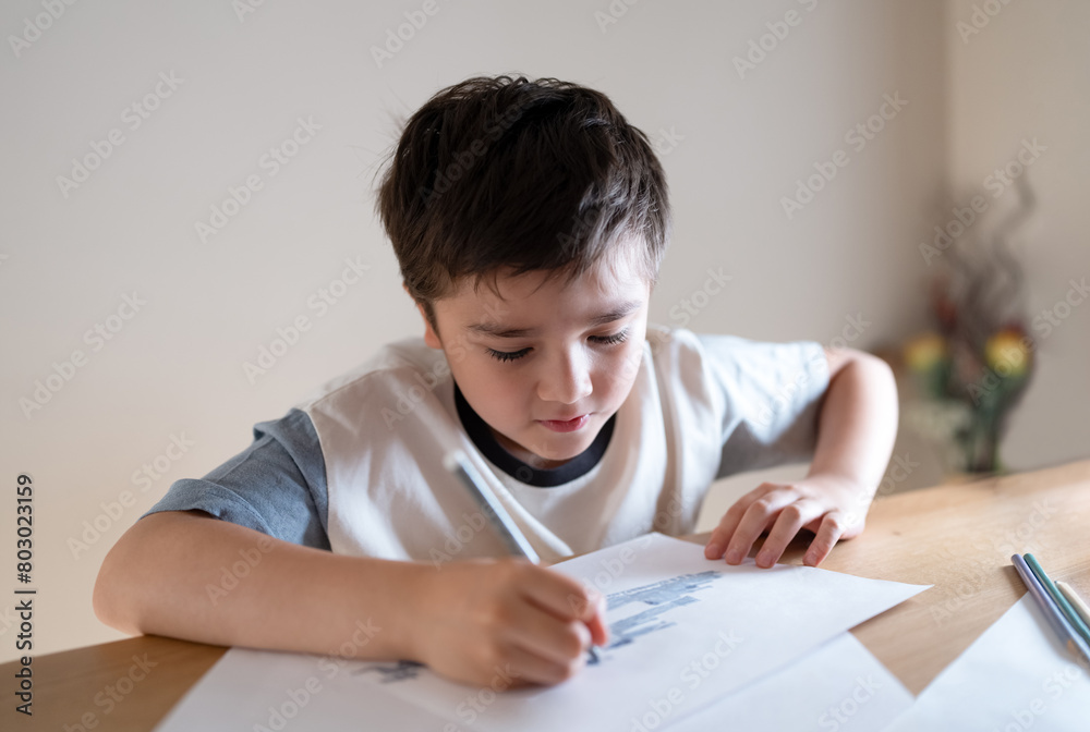 Kid siting on table doing homework,Child boy holding grey pen clouring and drawing on white paper, Elementary school and home schooling, Distance Education concept. Kid siting on table doing homework,Child boy holding grey pen clouring and drawing on white paper, Elementary school and home schooling, Distance Education concept.