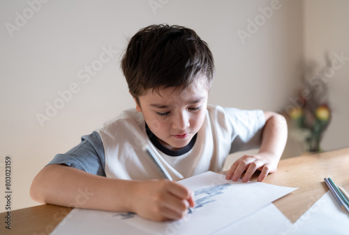 Kid siting on table doing homework,Child boy holding grey pen clouring and drawing on white paper, Elementary school and home schooling, Distance Education concept.
