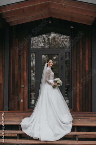 A woman in a white dress stands on a wooden staircase in front of a building. She is holding a bouquet of flowers and a veil. The scene is likely a wedding or a formal event