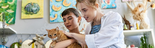 A loving lesbian couple at an art studio, with one woman gently holding a cat in her arms.