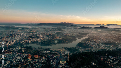 Aerial view of Dalat City at sunrise, featuring the tranquil Xuan Huong Lake amidst urban structures, surrounded by lush greenery and misty hills under a golden sky.