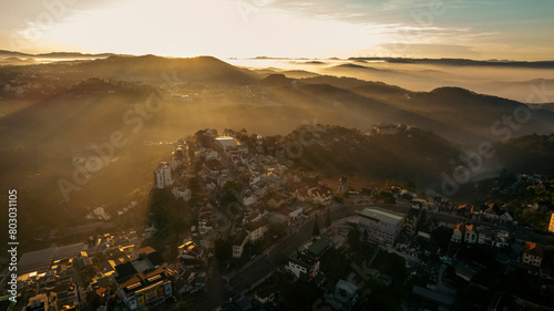 Aerial view of Da Lat at sunrise, showcasing mist-covered buildings amidst lush mountains, under a golden sky, highlighting the city’s architectural and natural beauty.