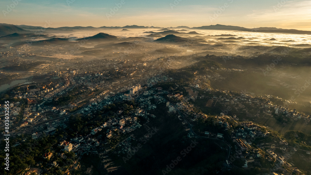 Aerial view of Da Lat at sunrise, showcasing mist-covered buildings amidst lush mountains, under a golden sky, highlighting the city’s architectural and natural beauty. Aerial view of Da Lat at sunrise, showcasing mist-covered buildings amidst lush mountains, under a golden sky, highlighting the city’s architectural and natural beauty.