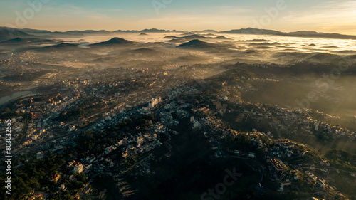 Aerial view of Da Lat at sunrise  showcasing mist-covered buildings amidst lush mountains  under a golden sky  highlighting the city   s architectural and natural beauty.