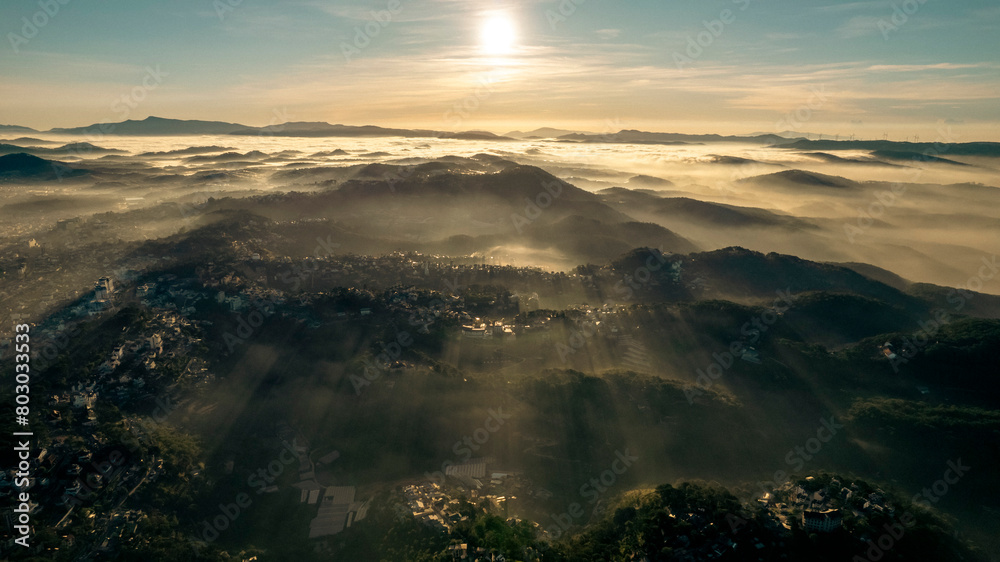 Aerial view of Da Lat at sunrise, showcasing mist-covered buildings amidst lush mountains, under a golden sky, highlighting the city’s architectural and natural beauty. Aerial view of Da Lat at sunrise, showcasing mist-covered buildings amidst lush mountains, under a golden sky, highlighting the city’s architectural and natural beauty.