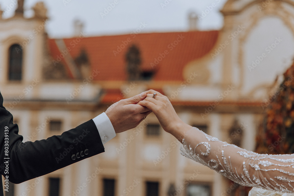 A couple is holding hands in front of a building. The man is wearing a wedding ring. The woman is wearing a long sleeved dress