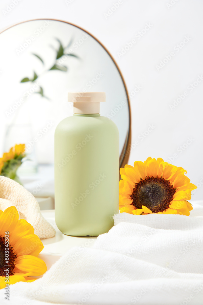 Blank green bottle stand on white table, surrounded by some bathroom products such as bath towel and mirror with sunflower as an accent. Space for design and front view