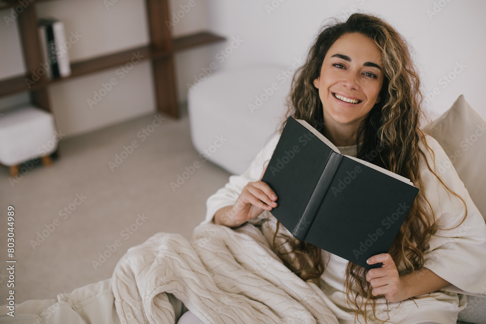 © polinaloves - Young woman reading book, sitting on sofa in a cozy living room.