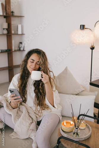 Young woman using her smartphone and drinking tea, relaxing in a cozy living room.