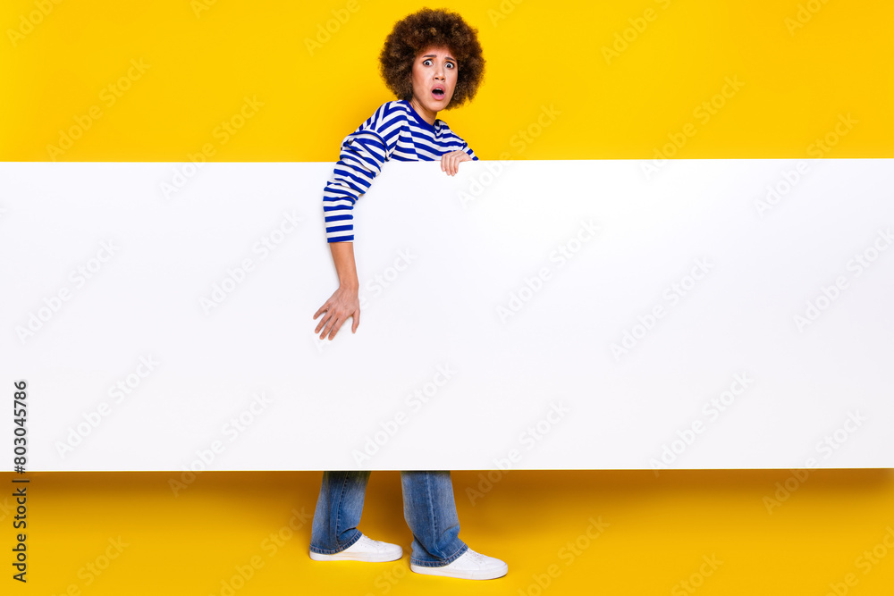 Full size photo of frightened girl wear striped pullover hold banner empty space astonished staring isolated on yellow color background Full size photo of frightened girl wear striped pullover hold banner empty space astonished staring isolated on yellow color background