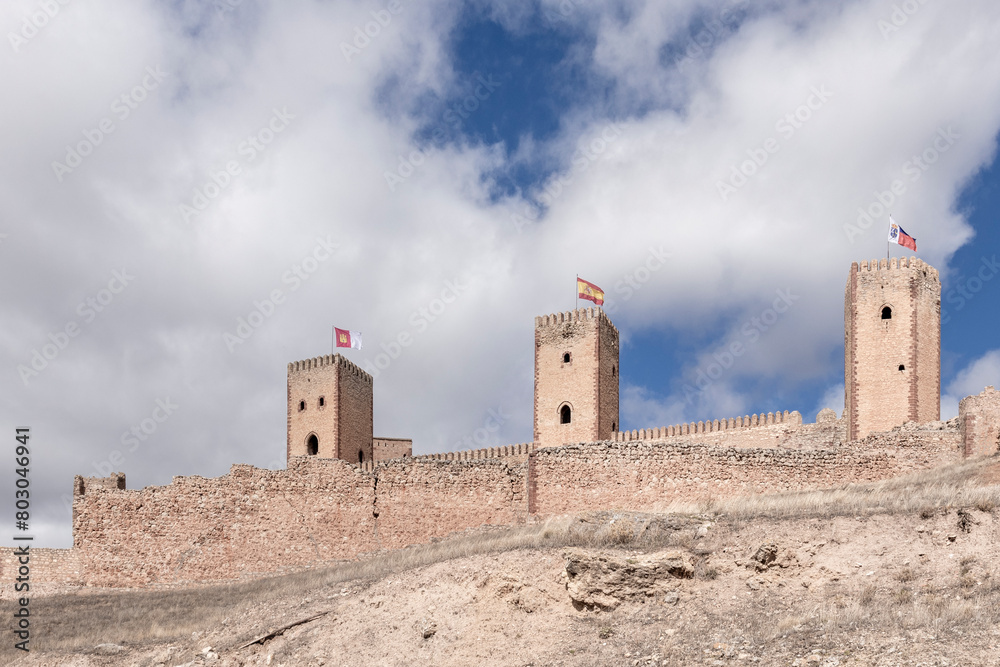 An ancient stone castle atop a hill, with three towers and flags, under a partly cloudy sky