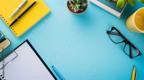 neat workspace with a yellow notebook, glasses, pens, a cactus, clipboard and a cup on a vibrant blue background. photo