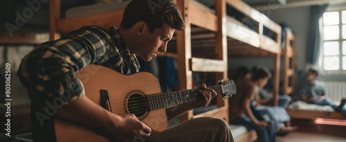 Young tourist sitting on bunk bed and playing a guitar in hostel room. Roommates singing and partying in contemporary youth hostel.