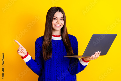 Photo portrait of pretty teen girl hold netbook point empty space wear trendy knitwear blue outfit isolated on yellow color background